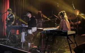 Dressed Like Boys, concert aux 3 Baudets, (MaMA 2025). Jelle Denturck au piano et synthétiseurs, entouré de son groupe, sous éclairage tamisé et rideaux rouges.