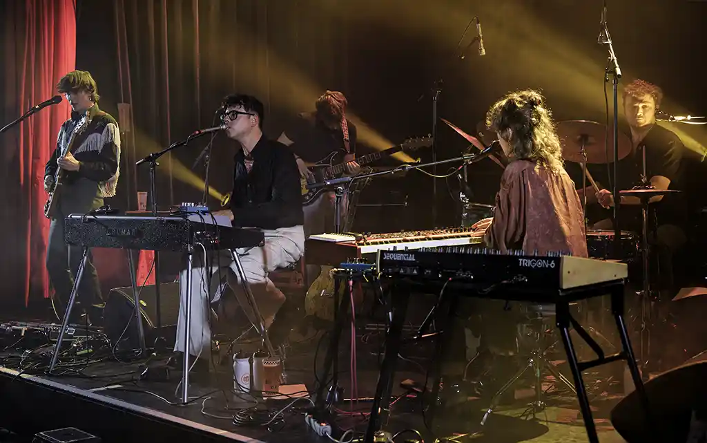 Dressed Like Boys, concert aux 3 Baudets, (MaMA 2025). Jelle Denturck au piano et synthétiseurs, entouré de son groupe, sous éclairage tamisé et rideaux rouges.